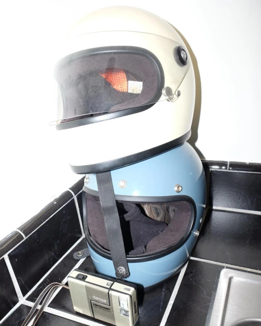 Stacked motorcycle helmets in white and blue, with a vintage camera resting on black-and-white tiled surface.