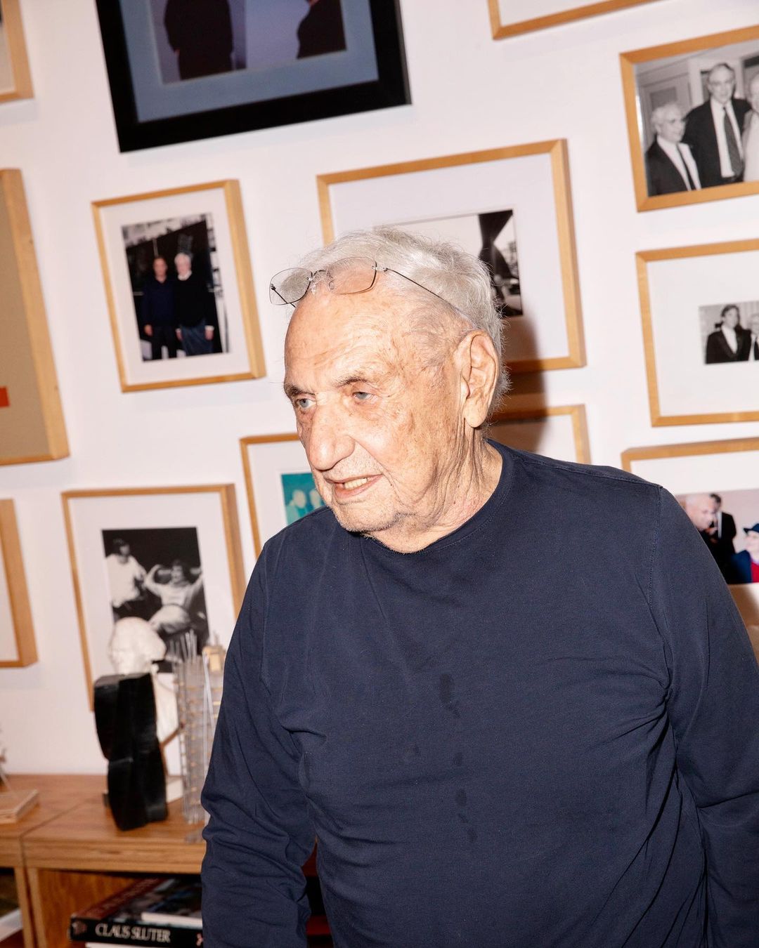 Older gentleman with glasses stands indoors wearing a navy top with a gallery wall of framed photos behind him in soft, natural light.