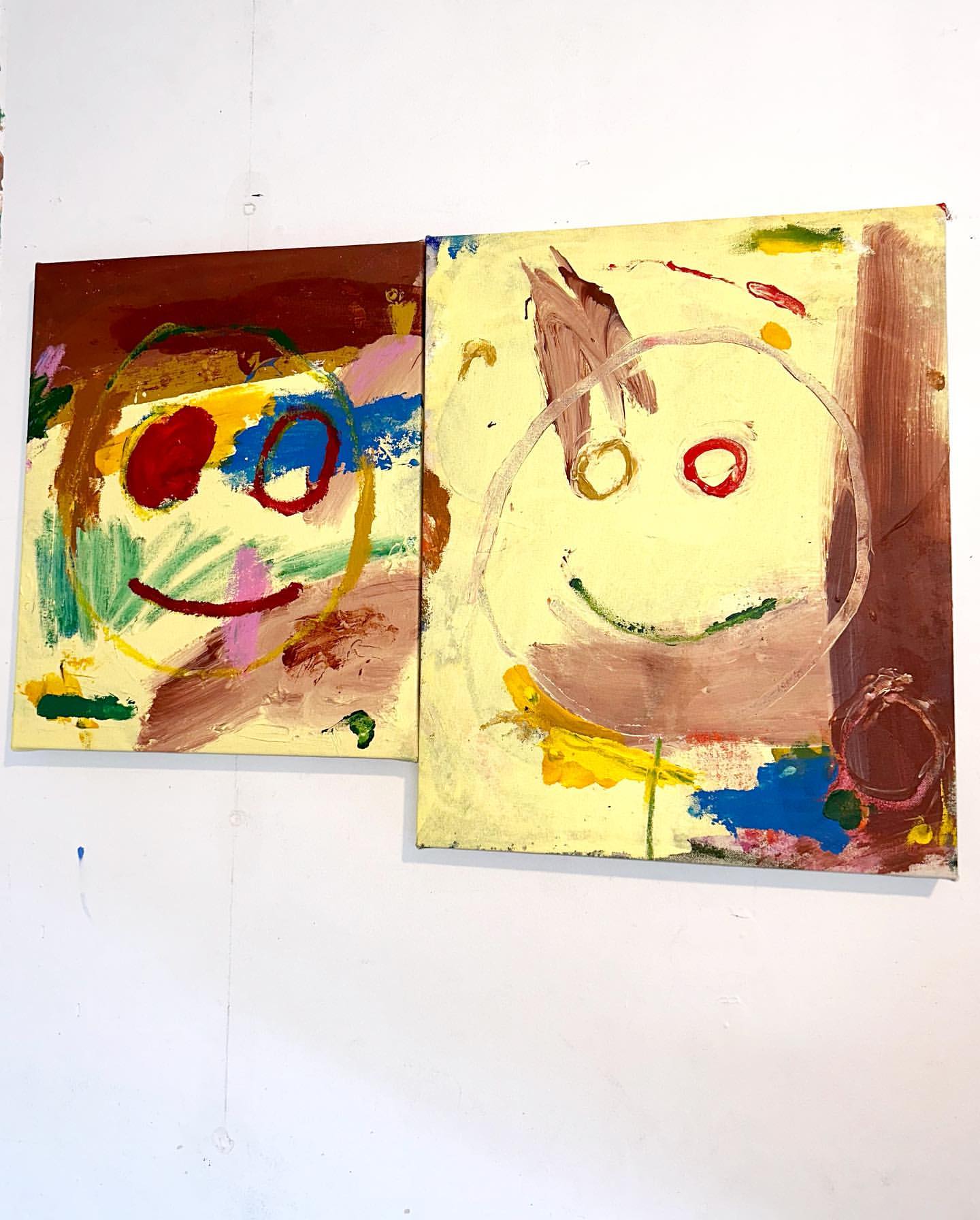 Two abstract, colorful, childlike paintings of smiley faces. One with a brown background and one with a yellow background, indoor studio shot.