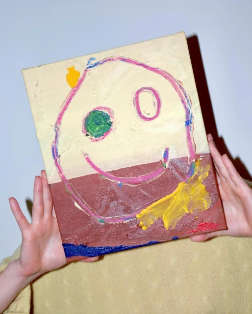 Child's art: Hands holding up canvas with painted smiley face in brown, yellow, blue, green, and pink against a plain background.