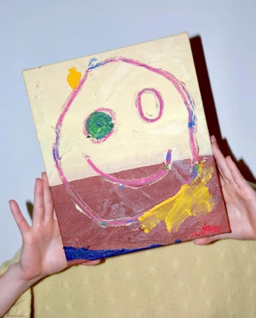 Child's art: Hands holding up canvas with painted smiley face in brown, yellow, blue, green, and pink against a plain background.