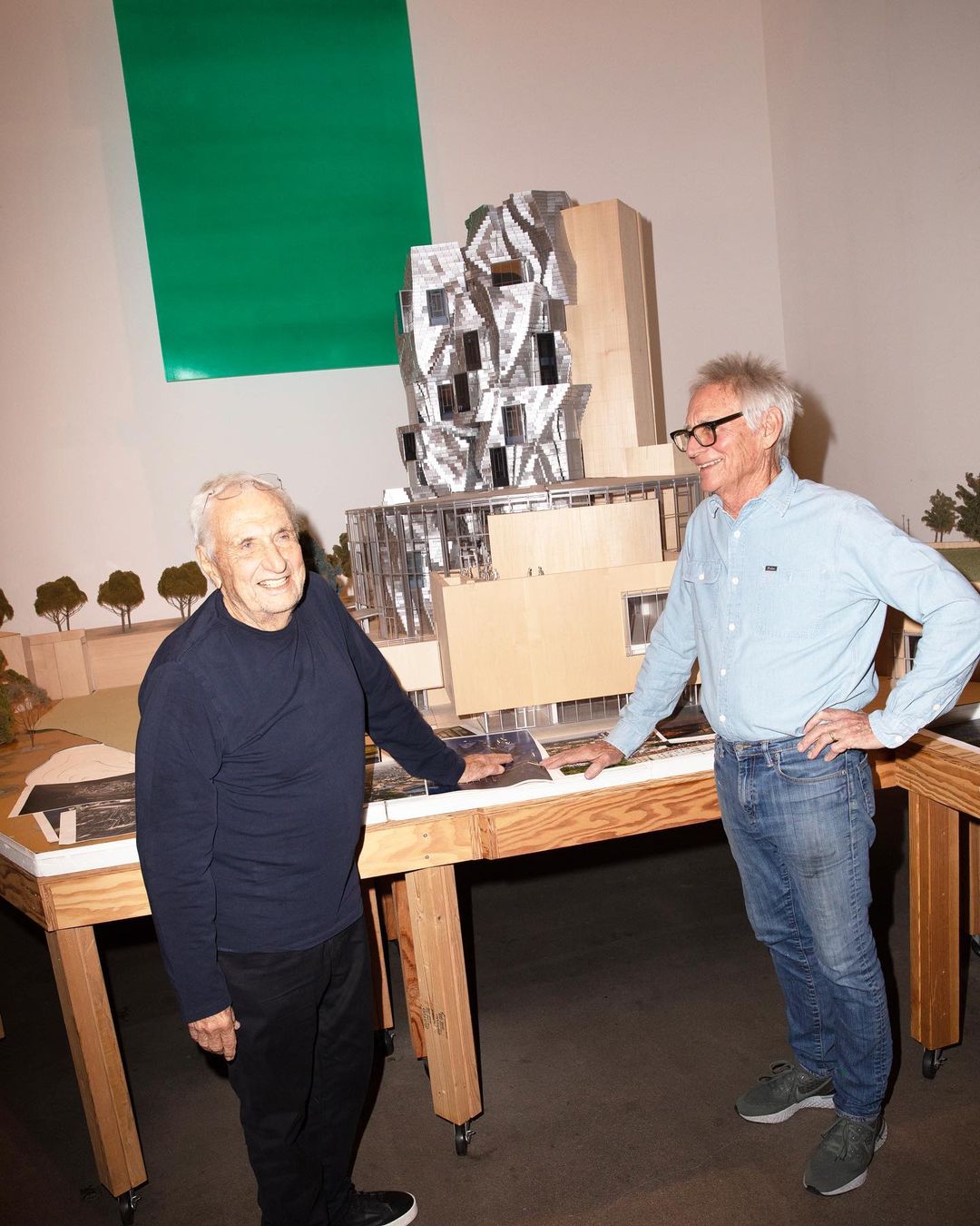 Two older men stand next to an architectural model of a building. One wears a dark sweater, the other a light blue shirt and jeans.