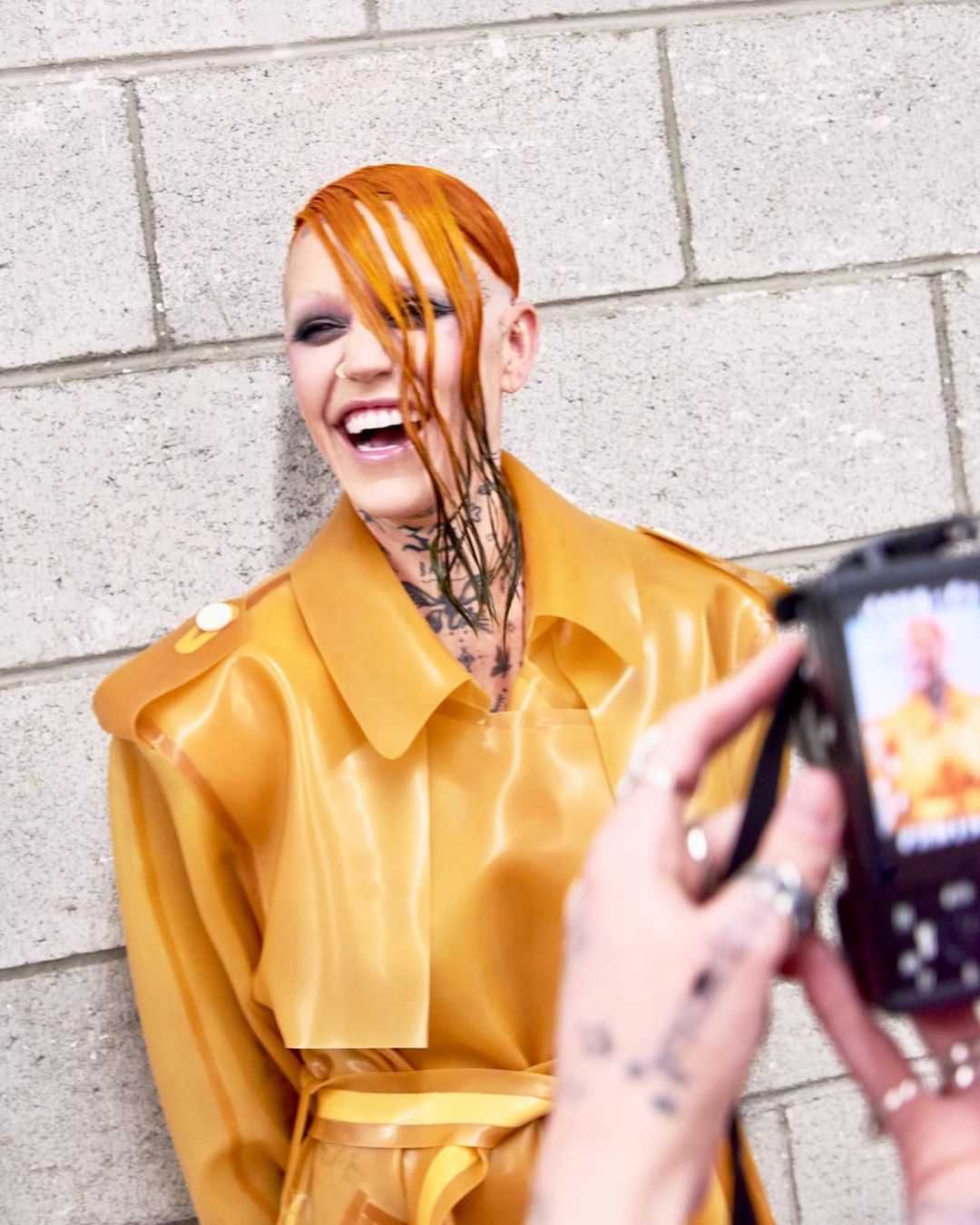 Person with orange hair and tattoos smiles while another person takes a photo with a digital camera against a gray brick wall.