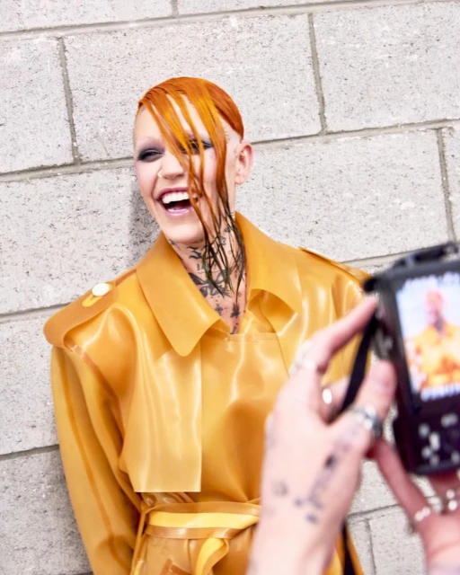 Person with orange hair and tattoos smiles while another person takes a photo with a digital camera against a gray brick wall.