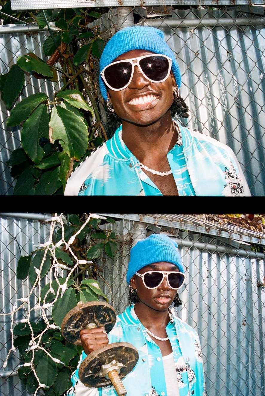 Athletic Black man smiling outdoors in beanie and sunglasses, wearing colorful jacket and necklace. He is lifting a dumbbell.