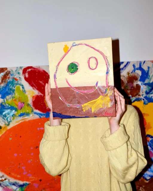 Person holding up a child-like painting of a smiling face with abstract painting visible in the background.
