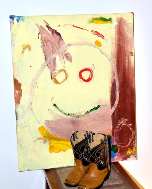 Whimsical art featuring an abstract smiley face painting paired with miniature cowboy boots. Modern art and western theme.