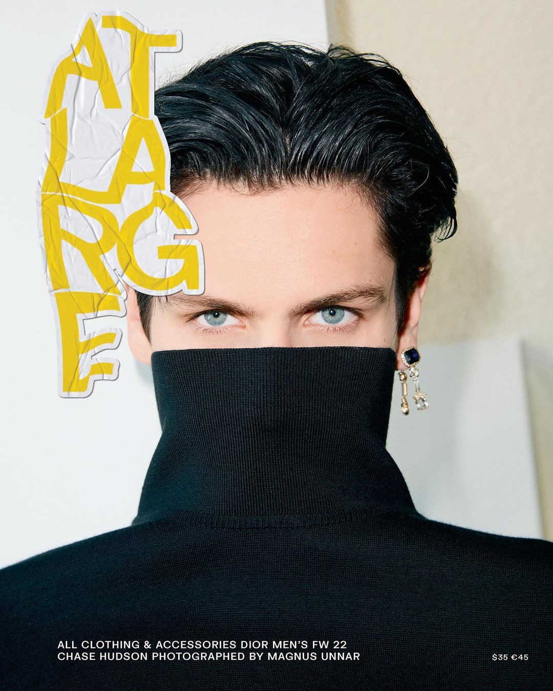 Portrait of a fair-skinned adult male in a black turtleneck with blue eyes, glossy black hair, and a black earring against a neutral backdrop.