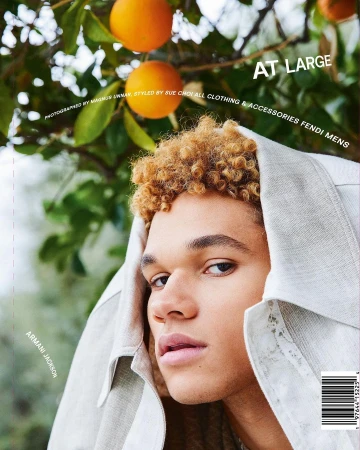 Close-up portrait of a young man with curly blonde hair wearing a hooded light garment under an orange tree with ripe fruit.