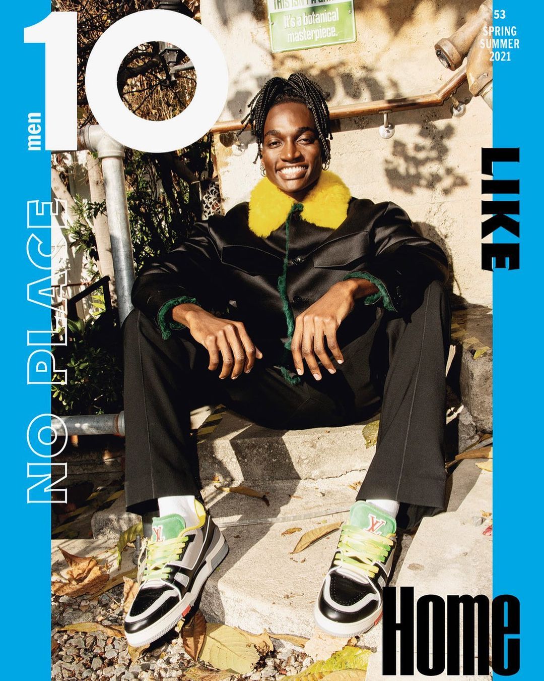 Magazine cover featuring a smiling Black man with locs wearing a jacket with yellow faux fur collar sitting on concrete steps.