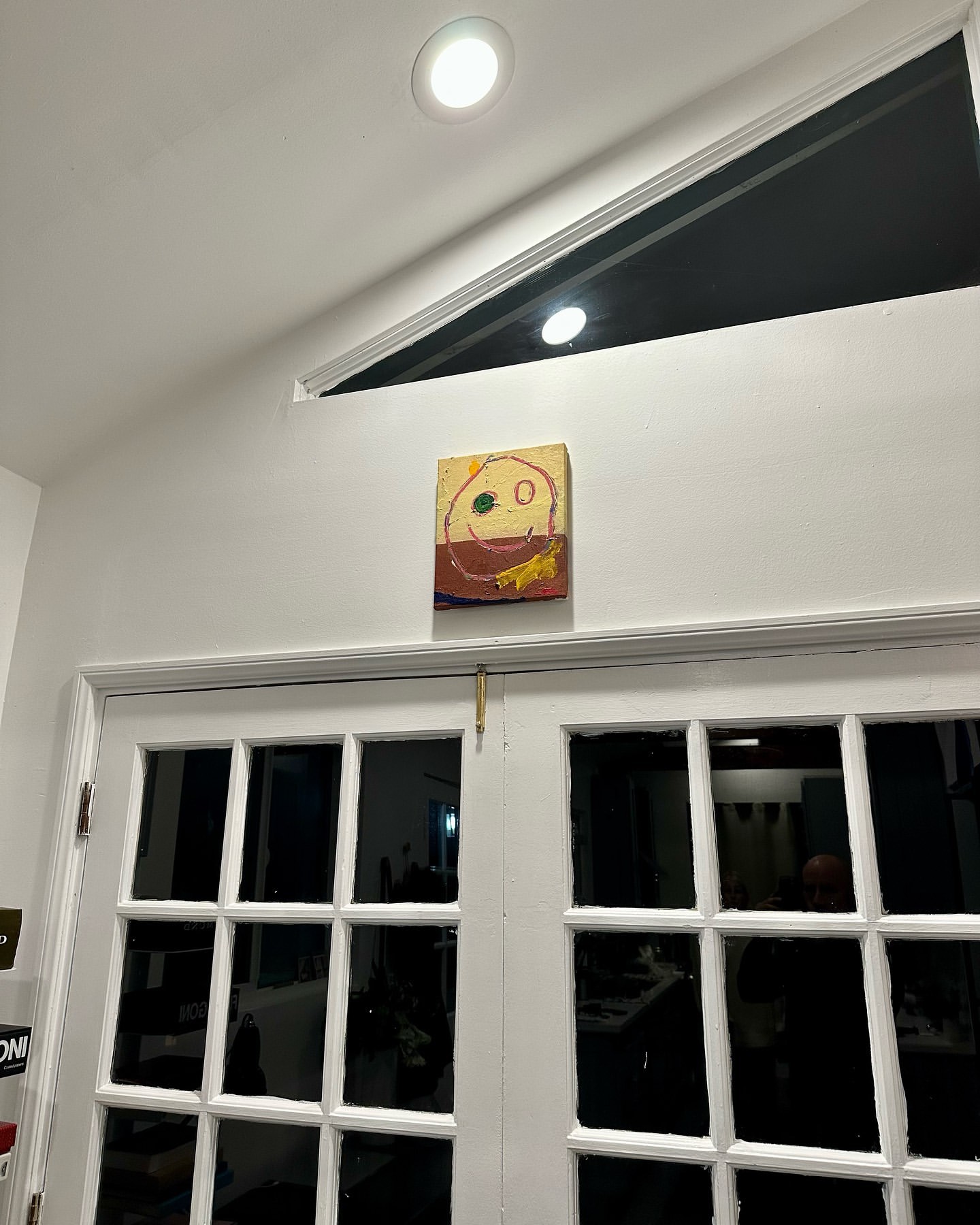 Interior shot of a white-walled room with white double doors and a painting of a smiley face hanging above the doors.