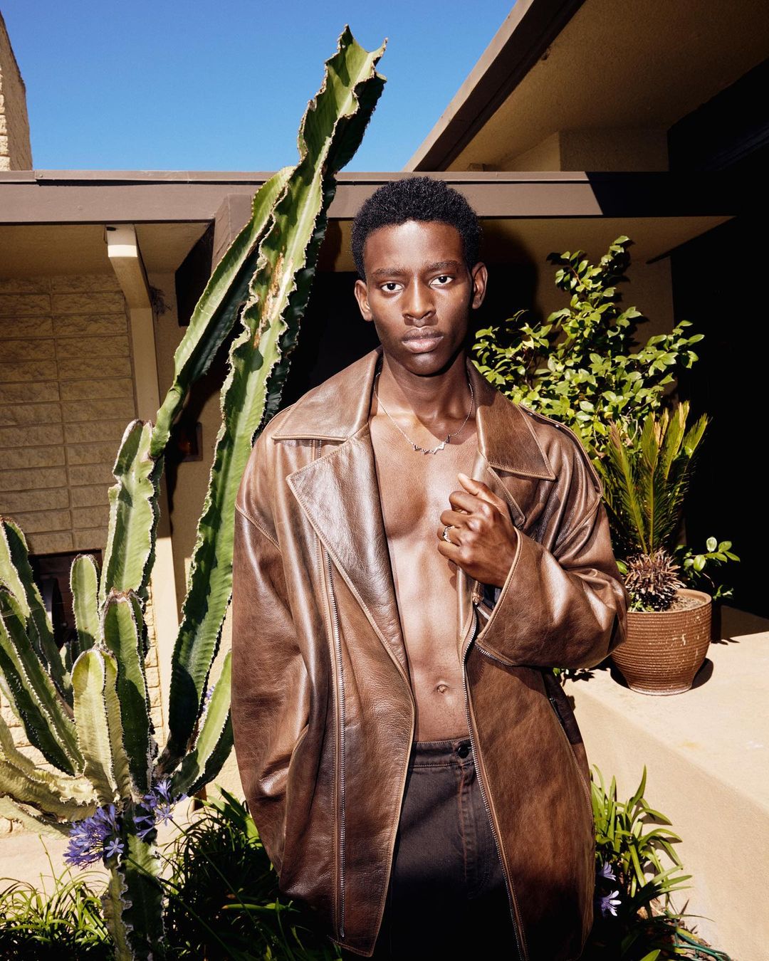 Portrait of a young Black man in a brown leather jacket standing outdoors with a cactus and building visible in the background.