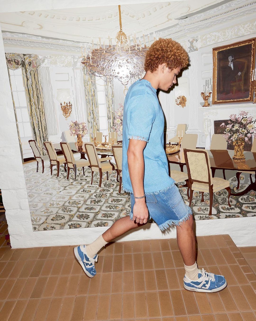 A young adult male in a blue shirt and jean shorts walks on a tile floor in front of a wall with a mural of a dining room.