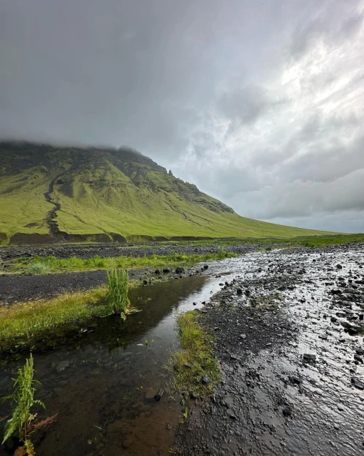 Landscape view of a green hillside under an overcast sky with a shallow stream and rocky foreground in an outdoor, natural setting.