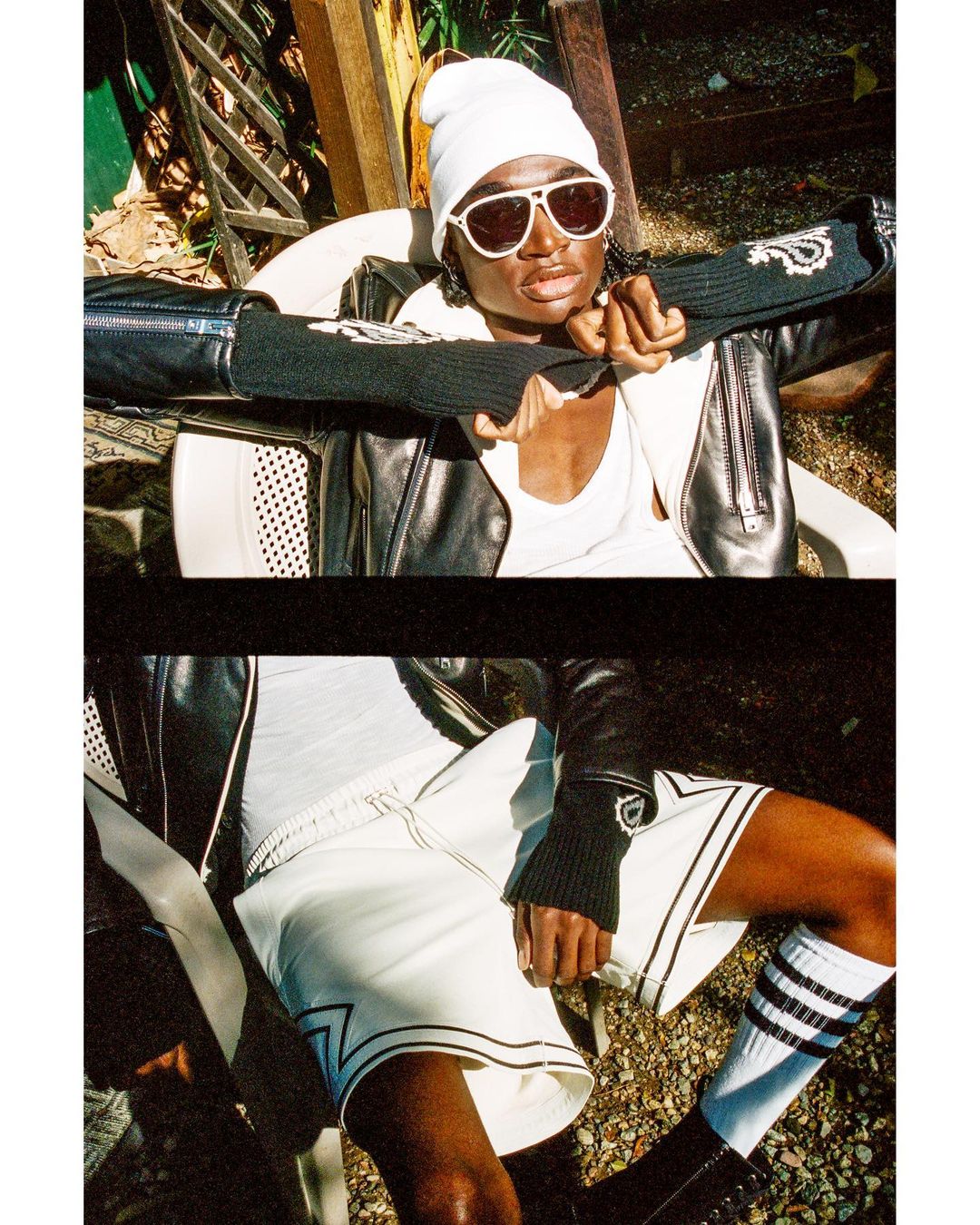 Outdoor shot of a black adult wearing sunglasses, white beanie, and leather jacket sitting in a chair with legs extended and crossed.