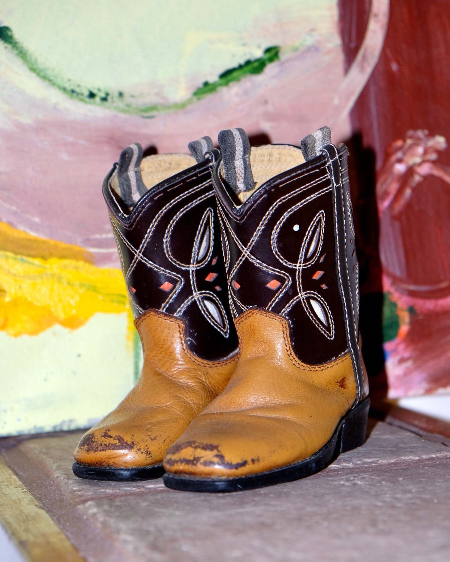 Two miniature tan and brown leather cowboy boots with stitching details sit side-by-side on a tan surface with a painted background.