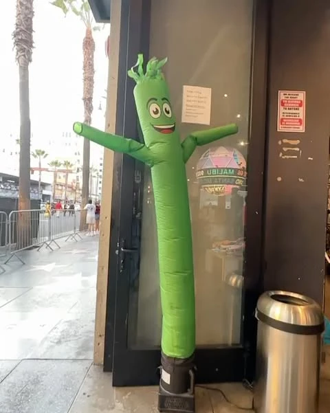 A green inflatable tube man stands outside a store, with palm trees and people visible in the background on a city street.