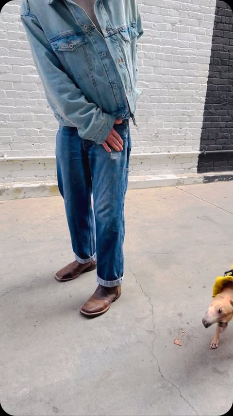 A person in a denim jacket and blue jeans stands with a dog in an outdoor, urban setting, capturing a casual, street-style aesthetic.