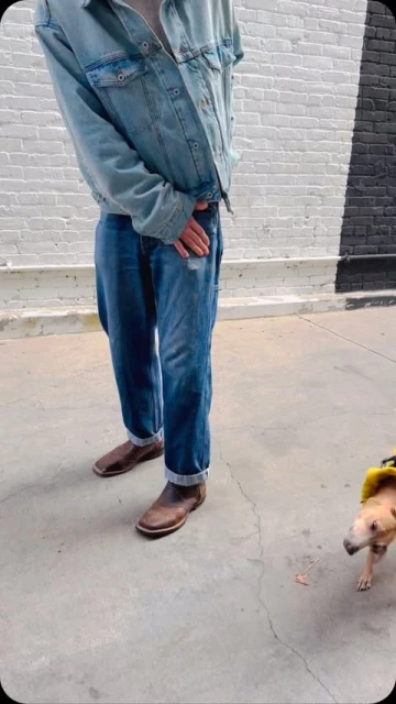 A person in a denim jacket and blue jeans stands with a dog in an outdoor, urban setting, capturing a casual, street-style aesthetic.