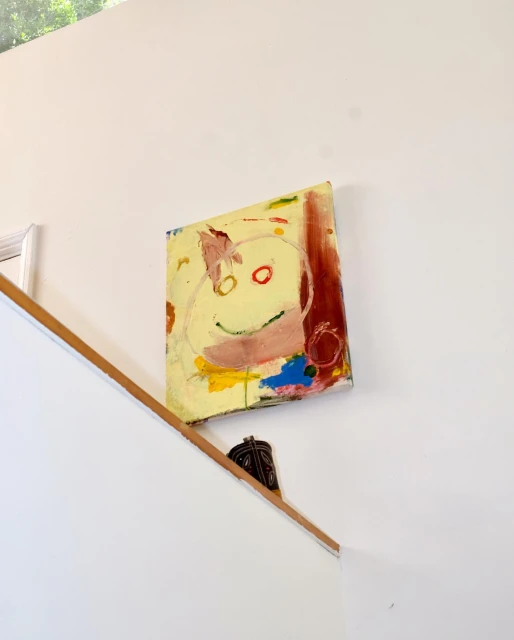 Abstract art painting featuring a cartoon face with red eyes and a pink mouth hanging on a white wall near wooden stairs.
