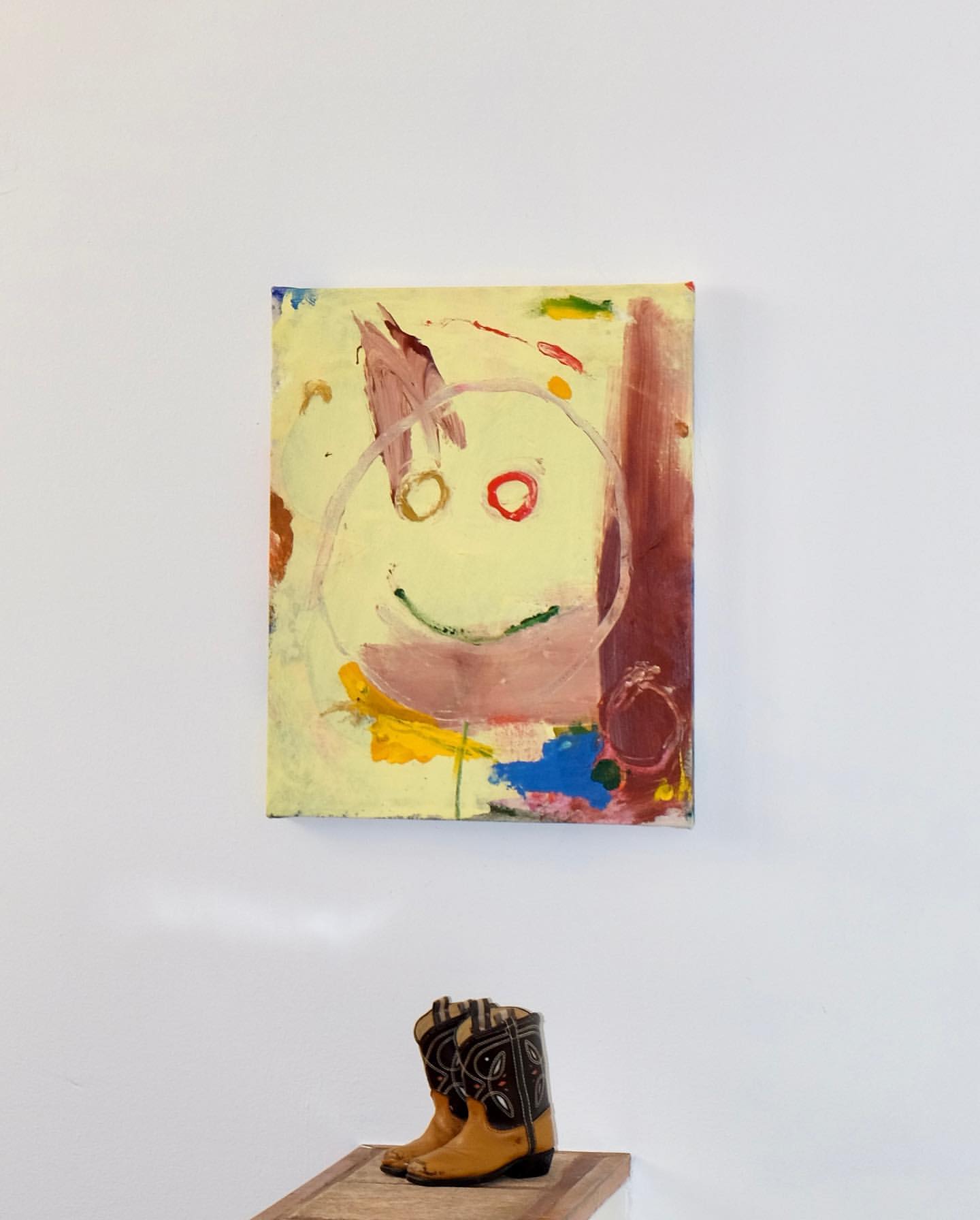 Abstract painting featuring a childlike face, hanging above a pair of western-style cowboy boots, creating a juxtaposition of art and fashion.