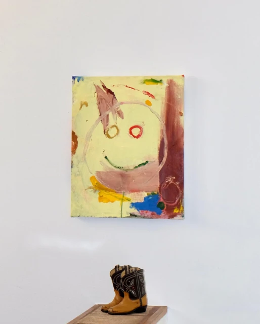 Abstract painting featuring a childlike face, hanging above a pair of western-style cowboy boots, creating a juxtaposition of art and fashion.