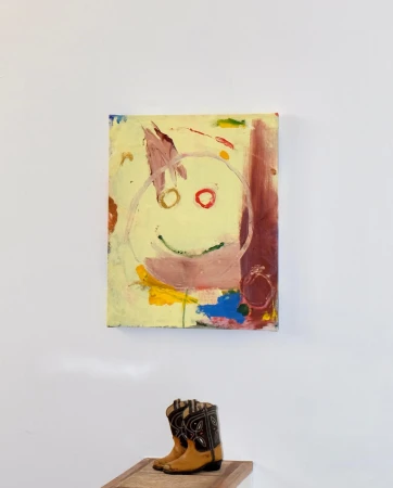Abstract painting featuring a childlike face, hanging above a pair of western-style cowboy boots, creating a juxtaposition of art and fashion.