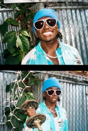 Portrait of a smiling Black man in a blue beanie and white sunglasses holding a rusty dumbbell against a chain link fence.