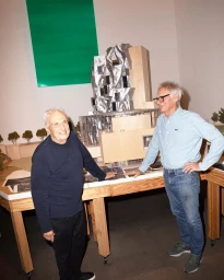 Two older men standing next to architectural models on tables with a green rectangle on the wall.