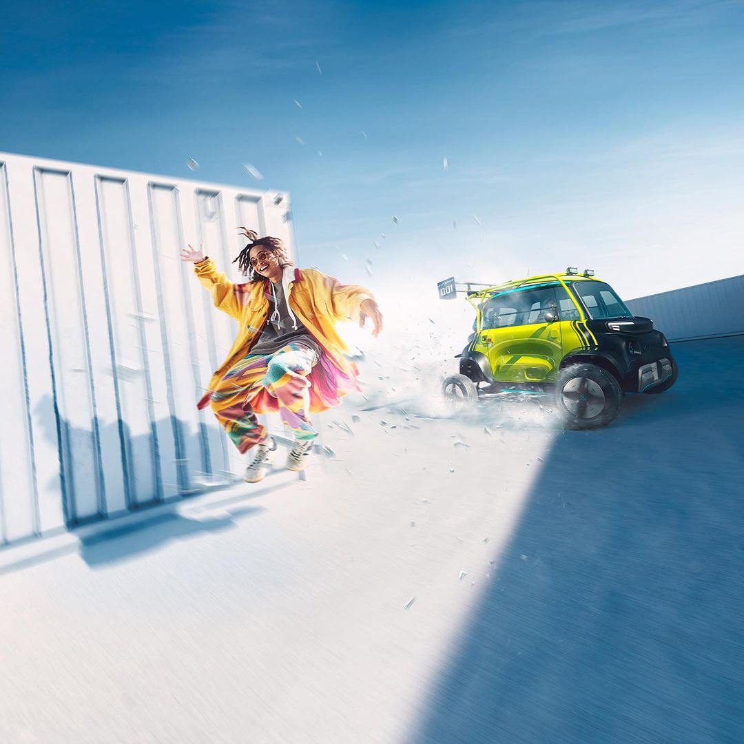 Person skateboarding near container as a small neon green car kicks up snow or dust, set against a blue and white sky backdrop.