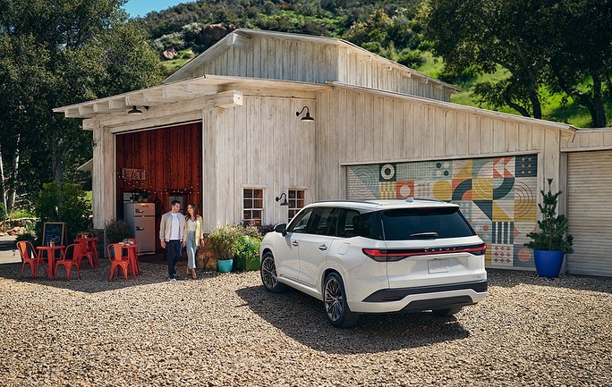 White SUV parked near a couple outside a white barn-style building with geometric art; rural setting with green hills in the background.