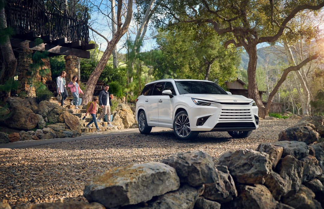 White SUV parked on a gravel path with family walking down stone stairs in the background, surrounded by trees and natural landscaping.