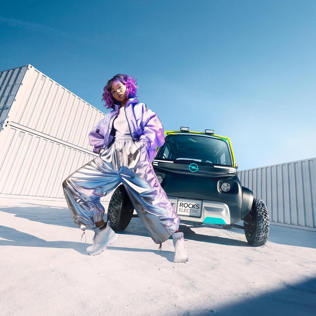 Young woman in a futuristic outfit poses next to a compact electric car, standing on a concrete surface with shipping containers in the back.