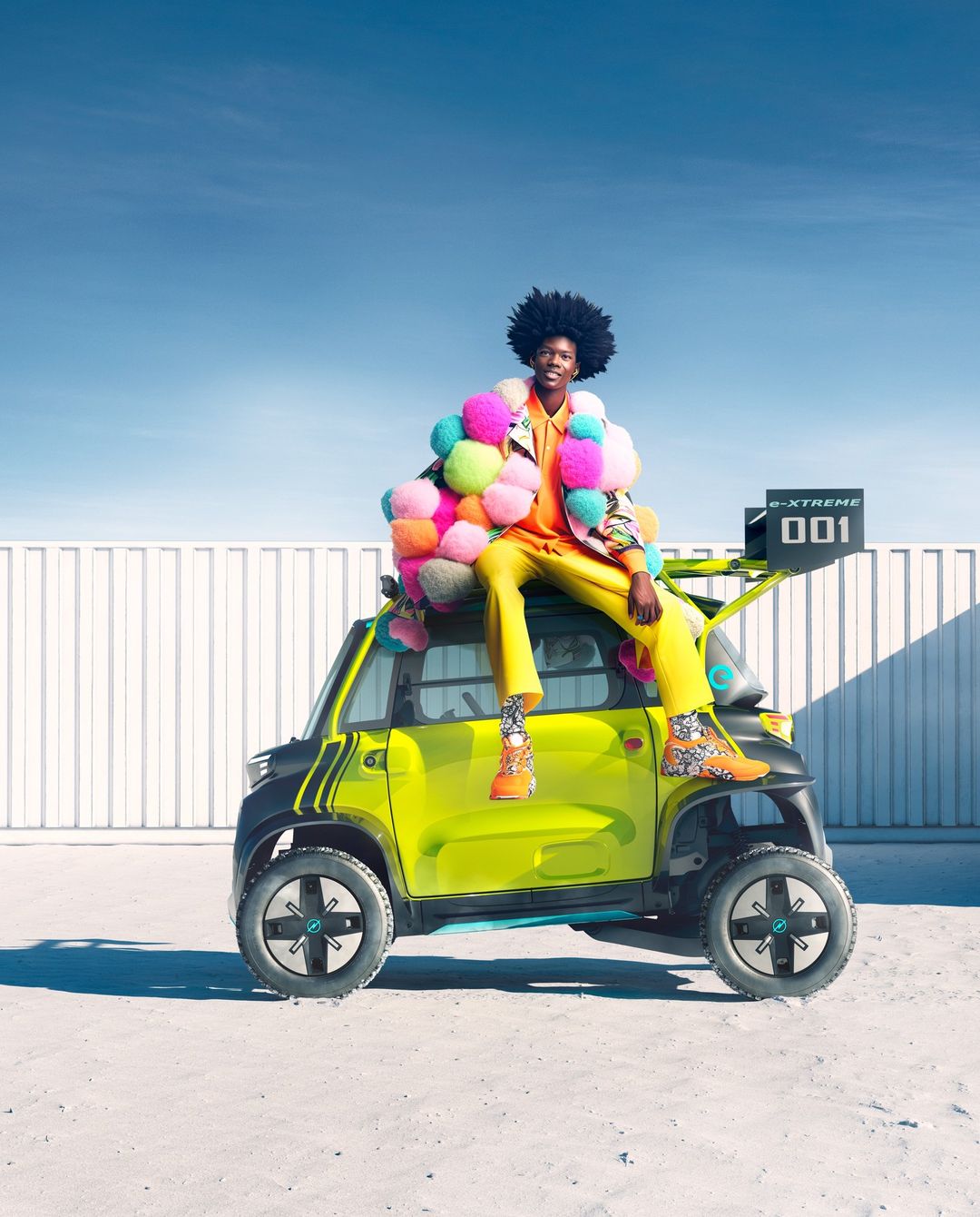 Stylish adult poses on a vibrant electric car against a blue sky, showcasing colorful, eye-catching fashion and automotive design.