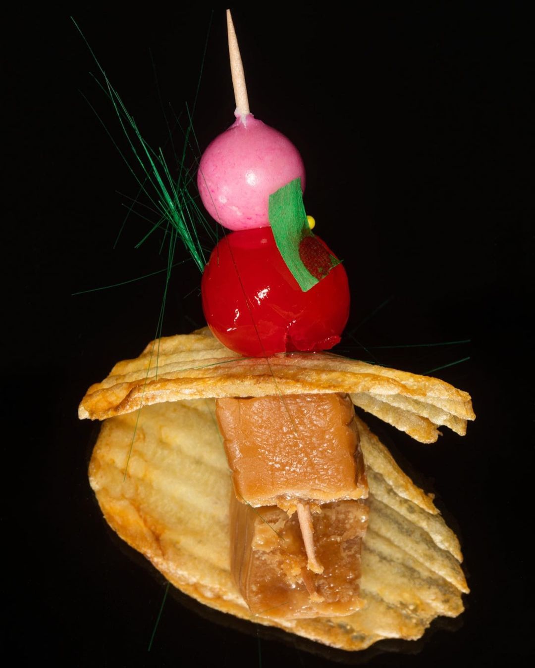 Colorful food skewer on a potato chip, featuring stacked gummy candy, a red glazed cherry, and a pink ball on a black background.