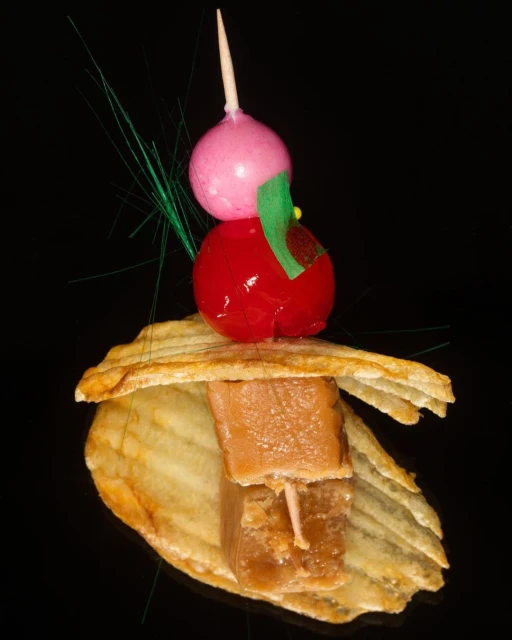 Colorful food skewer on a potato chip, featuring stacked gummy candy, a red glazed cherry, and a pink ball on a black background.
