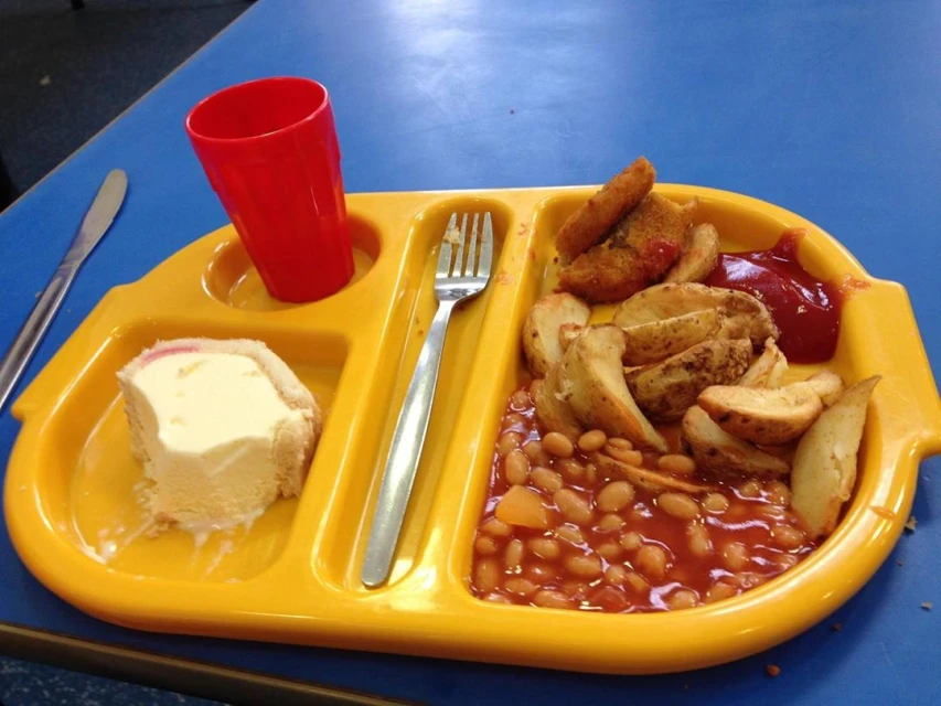 Yellow tray filled with baked beans, potato wedges, fish sticks, a red cup, and an ice cream dessert sitting on a blue table.