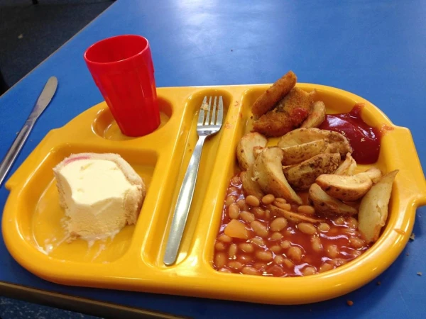 Yellow tray filled with baked beans, potato wedges, fish sticks, a red cup, and an ice cream dessert sitting on a blue table.