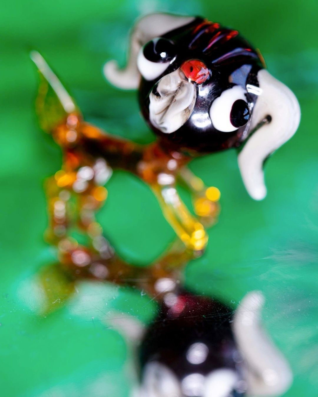 Close-up of a glass figurine of a ram with black head, white horns, and amber and brown body against a green reflective surface.