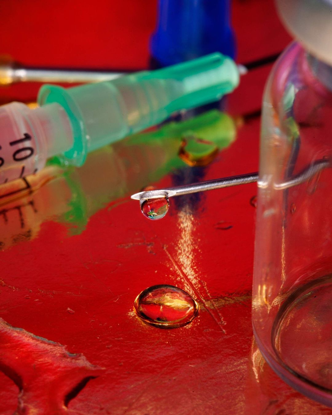 Close-up studio shot of a syringe with a needle and a vial on a red surface, showcasing a drop of liquid and medical equipment.