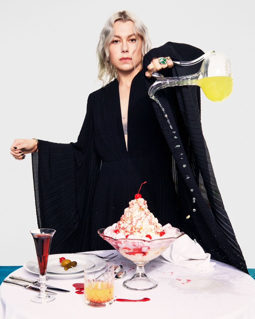 Woman in long black dress poses behind table with sundae and drinks, pouring liquid from glass pitcher in a studio setting.
