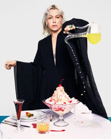 Woman in long black dress poses behind table with sundae and drinks, pouring liquid from glass pitcher in a studio setting.