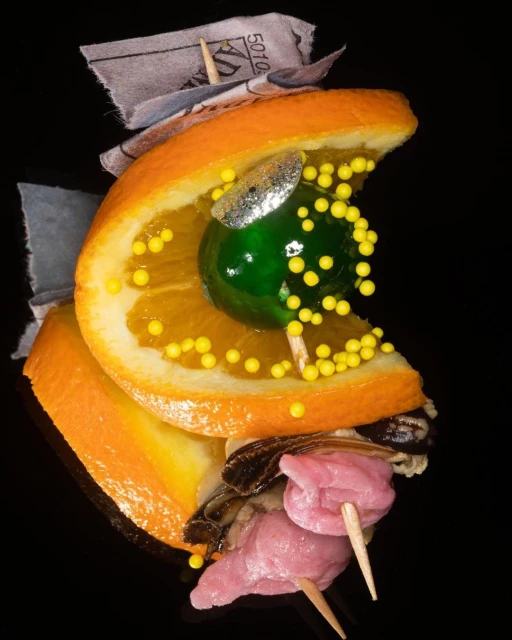 Surreal still life food art featuring skewered orange slices, green cherry, yellow pearls, oysters, and processed meat on a black backdrop.