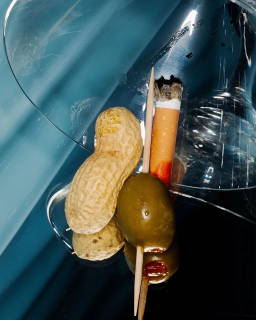 Close-up of a martini glass with peanuts, an olive skewer, and a cigarette against a blue gradient background in a studio setting.