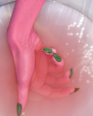 Close-up of a pink painted arm and hand with green nails, partially submerged in a white liquid inside a white container.