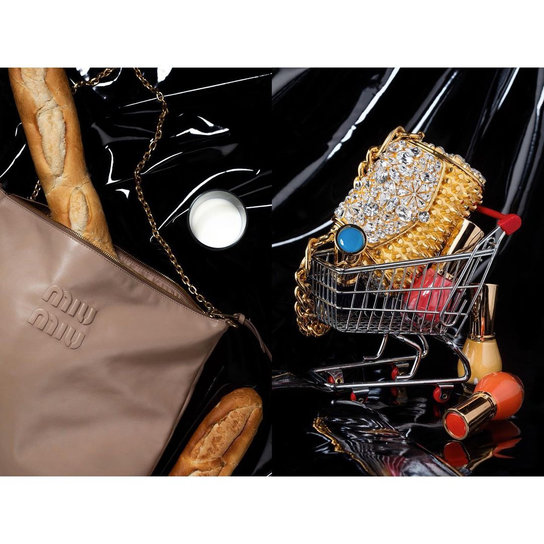 Still life featuring a Miu Miu bag, baguette, a jeweled purse in a miniature shopping cart, and makeup against a black background.