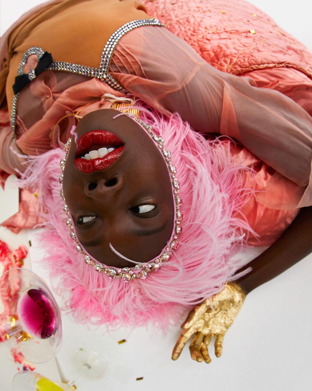 Elegant African American woman with pink feathered headpiece and gold hand poses with spilled wine and confetti on a white background.