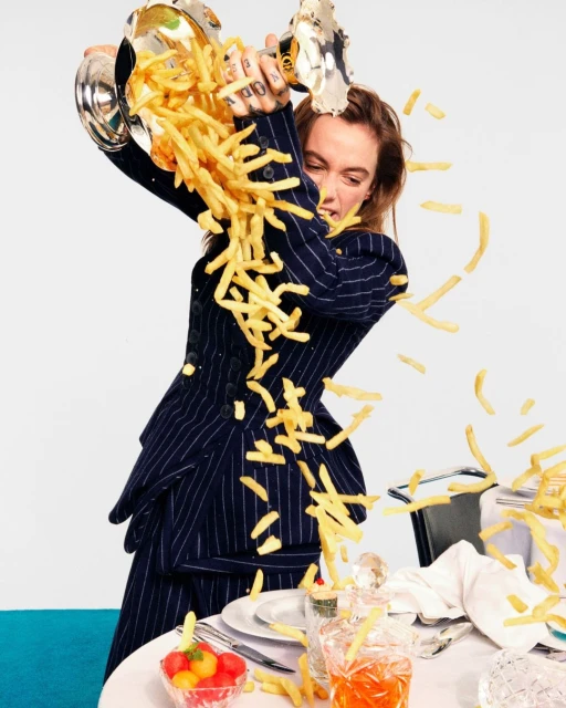 Woman in a pinstripe suit stands behind a set table, tossing fries into the air from a silver serving dish in a studio.