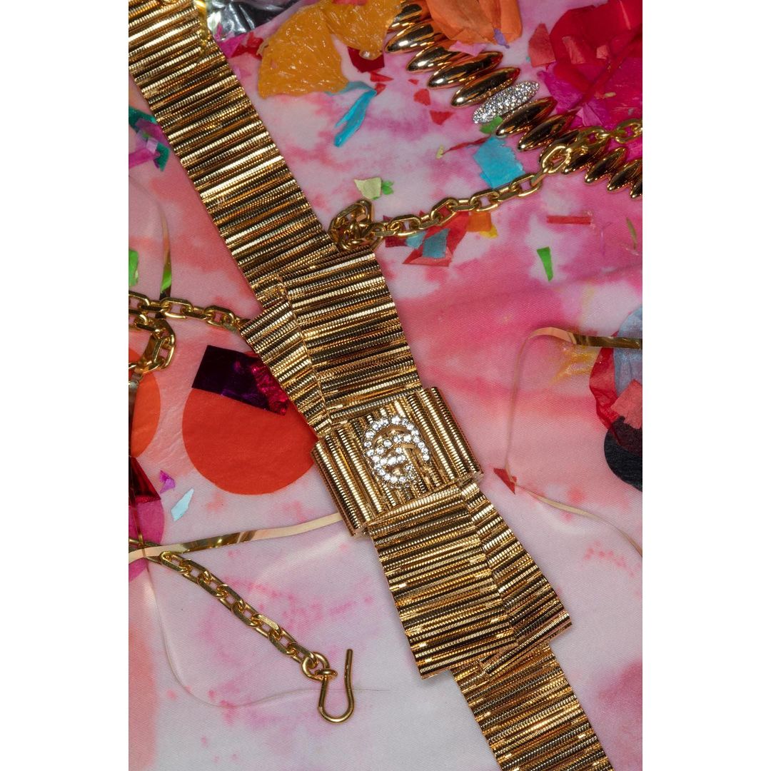Gold-toned chain belt with rhinestone-studded logo on a patterned background with confetti. Fashionable accessory for women's wear.