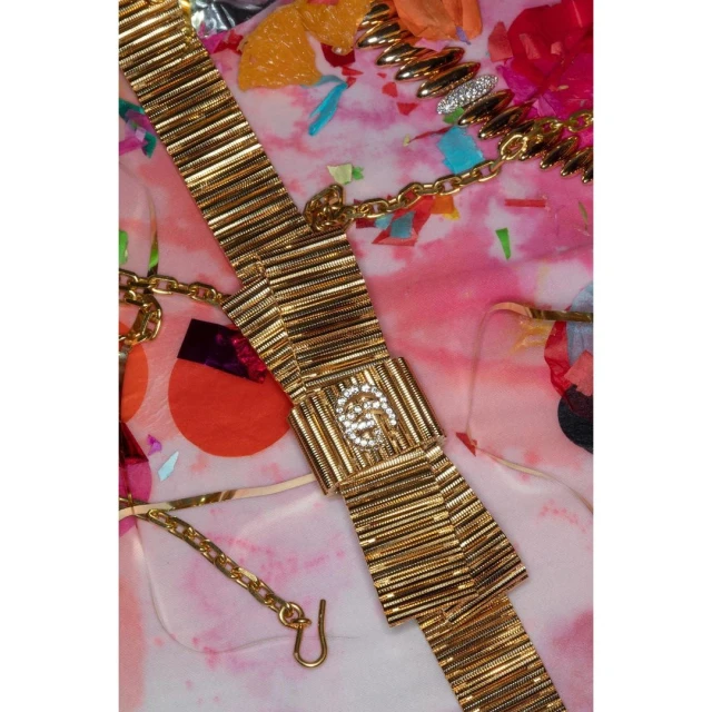 Gold-toned chain belt with rhinestone-studded logo on a patterned background with confetti. Fashionable accessory for women's wear.